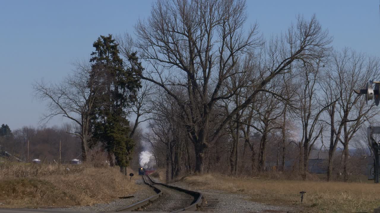 Long view of a restored steam locomotive and passenger cars traveling blowing smoke and steam following ahead of train thru farm countryside