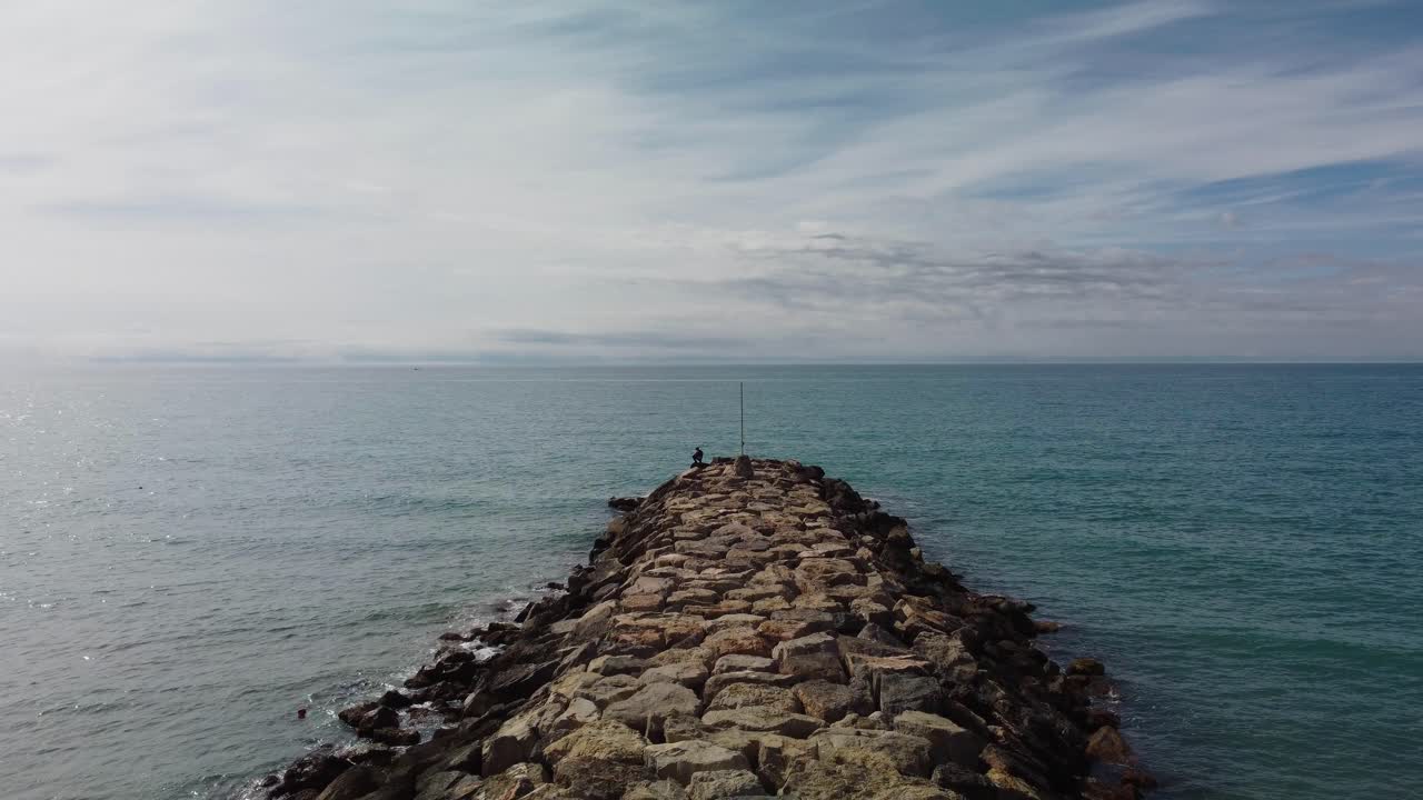 Rocky pier extending into the calm blue sea under a partly cloudy sky in Sitges, Spain