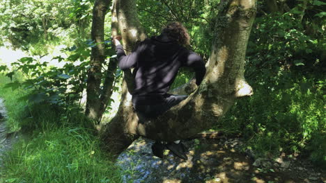 Person-Sitting-In-The-Tree-Over-The-Shallow-River-At-Summer