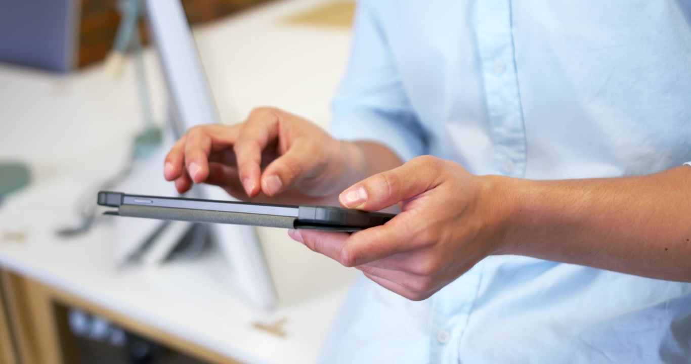 Man in blue shirt holding tablet case tapping top-left while swiping screen at desk, browsing app