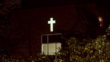 slow zoom on illuminated cross above window of old brick house; trees and bushes in foreground; eerie mood