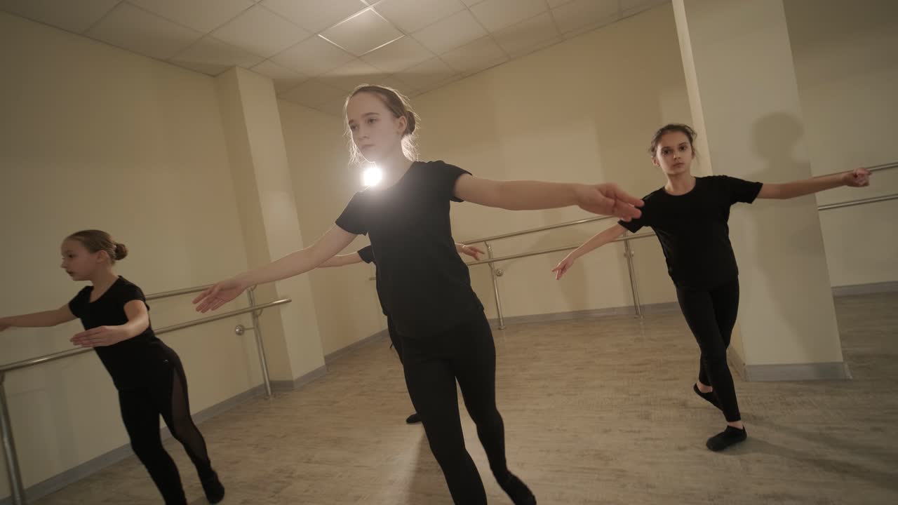 un grupo de jóvenes estudiantes de ballet con ropa de baile negra practicando posiciones en un amplio estudio de ballet con pisos de madera y barras montadas en la pared. expresiones enfocadas y movimientos sincronizados.