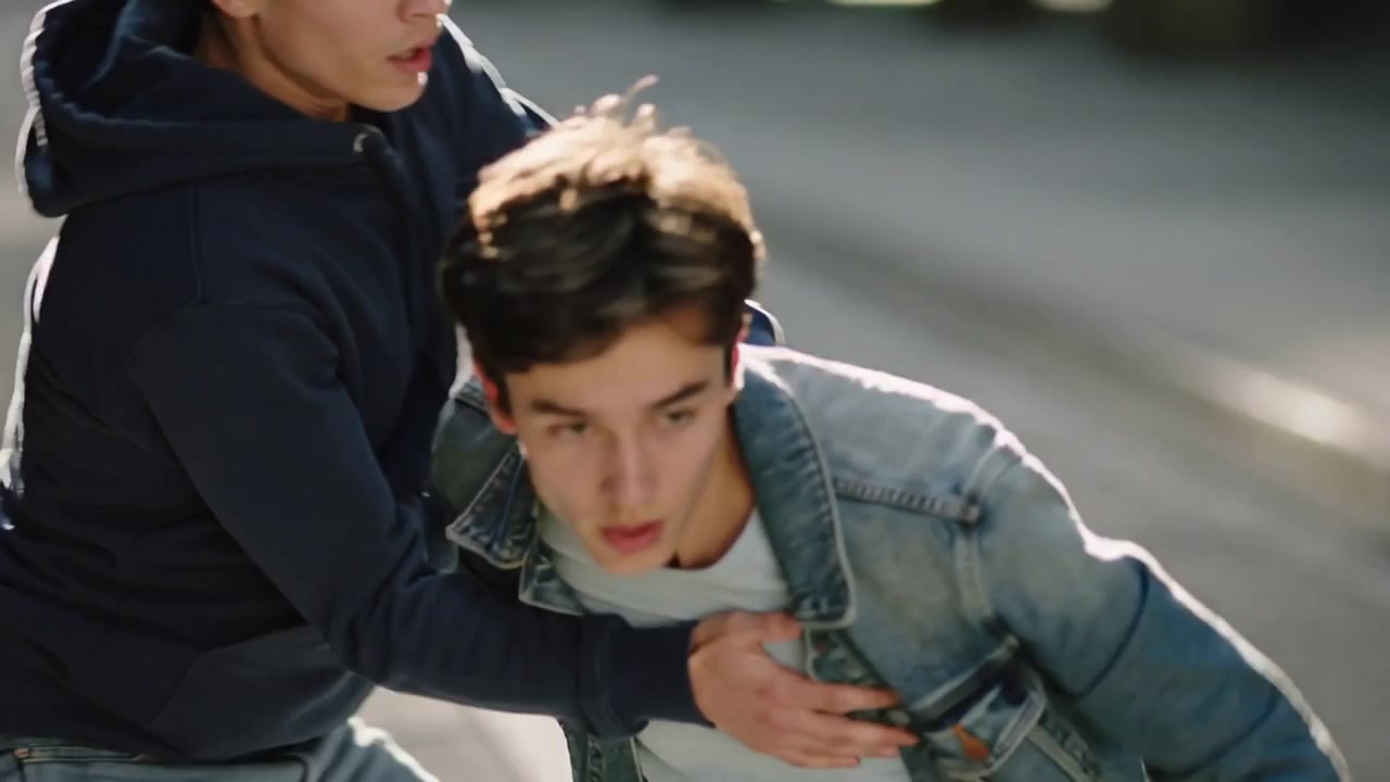 Adolescente in giacca di jeans che avanza, mentre una felpa blu navy si protende e lo aggancia per rallentarlo su una strada assolata
