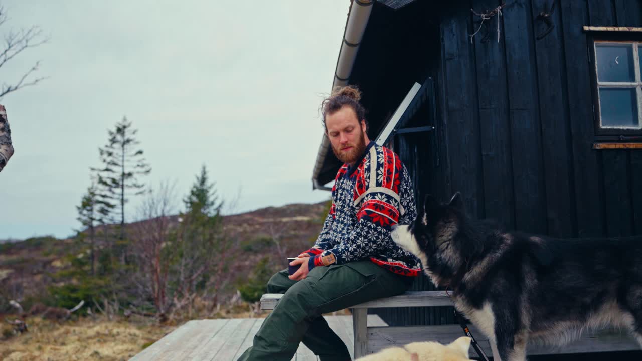 Norwegian Guy With Alaskan Malamute Dogs Resting Outside The Mountain Lodge. Static Shot