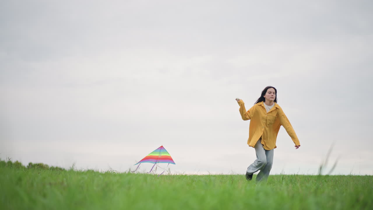 Mujer corriendo con una cometa en un campo, chaqueta amarilla ondeando bajo un cielo nublado. Una toma cinematográfica amplia captura la figura solitaria corriendo a través de una pradera verde, con el pelo ondeando al viento, transmitiendo una energía alegre y despreocupada.