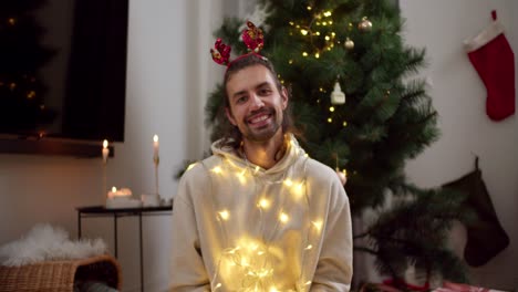 Portrait-of-a-happy-brunette-guy-in-a-white-sweater-who-looks-at-the-camera-and-smiles-in-a-New-Year's-yellow-glowing-garland-and-a-red-hoop-after-a-green-Christmas-tree-in-a-cozy-room-in-the-evening-in-winter