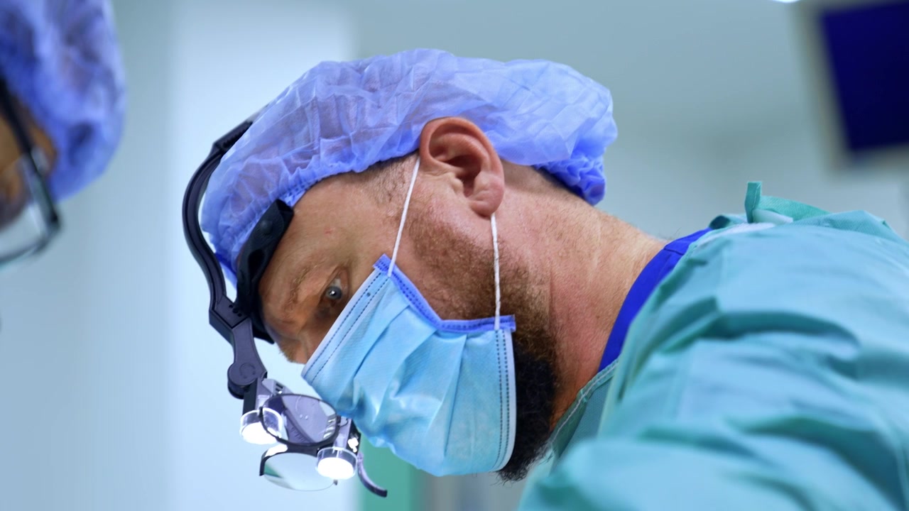 Medico maturo con la barba, con mascherina, cuffia e occhiali ingranditori chirurgici, lavora in sala operatoria. Ritratto di un chirurgo concentrato che esegue un'operazione. Primo piano.