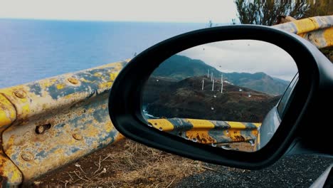 Car-side-mirror-reflection-of-countryside-vertical-wind-turbines-on-top-of-mountain-inside-parked-vehicle-green-energy-global-warming-windy-cloudy-day-tecnology-veiw-from-car-window