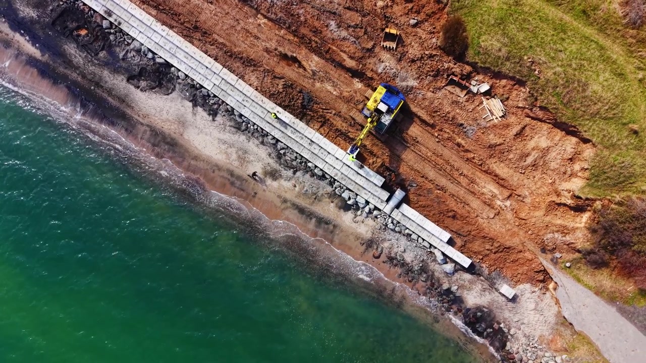 Vista aerea di un frangiflutti completato che incontra una spiaggia sabbiosa. Ripresa aerea con drone di un muro di cemento, mare turchese ed escavatore che continua la costruzione