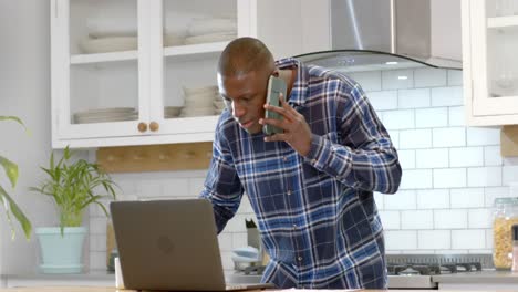 African-american-man-using-laptop-and-talking-on-smartphone-in-kitchen-at-home,-slow-motion