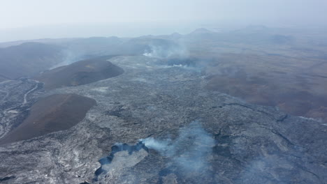 Otherworldly-aerial-drone-view-of-smoky-volcanic-lake-lava-landscape,-drone-flying-backwards-reveals-Fagradalsfjall-crater-cone,-Iceland,-sunny-day