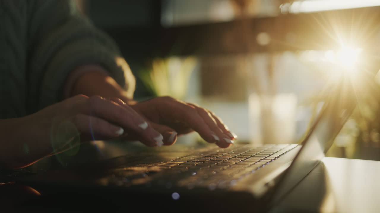 The hands of a middle-aged woman are typing text on a laptop keyboard. The setting sun illuminates her