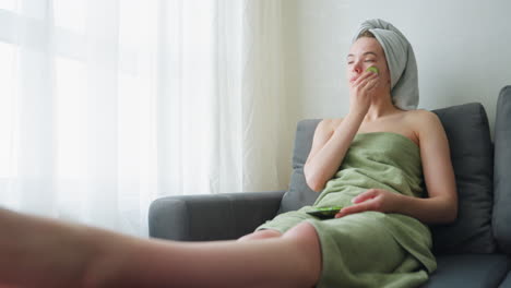 young girl sitting on couch wrapped in towel, gently rubbing cucumber on her face for skincare in a calm, bright indoor setting with natural light filtering through the window