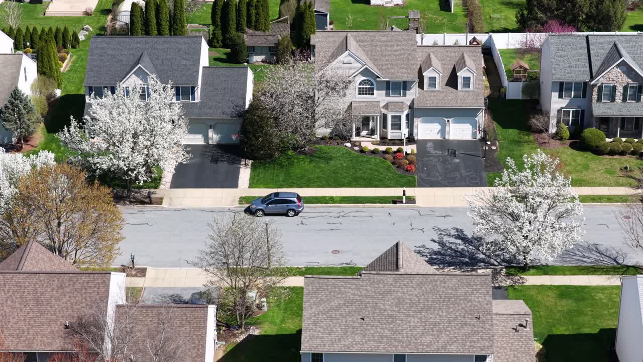 Vista aérea lateral de drone de um bairro suburbano tranquilo na Pensilvânia, EUA, com casas em estilo colonial, garagens, carros estacionados e árvores floridas na primavera em um ambiente residencial calmo. Plano geral.