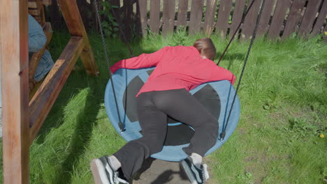 woman in red shirt lays face down on circular swing while adjusting position, enjoying sunny day in grassy backyard, another person partially visible swinging nearby