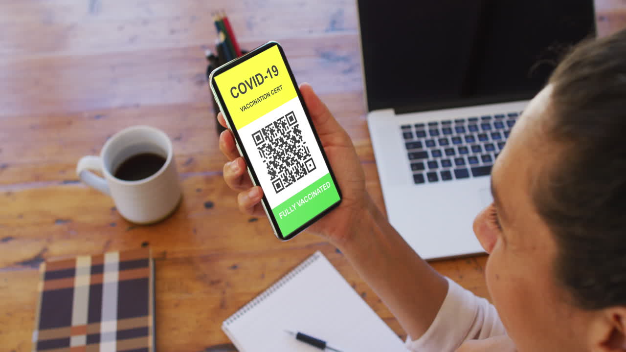 Woman at desk holding smartphone with covid vaccination certificate and qr code on screen