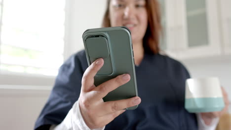 Happy-plus-size-biracial-female-doctor-having-coffee-and-using-smartphone-at-home,-slow-motion