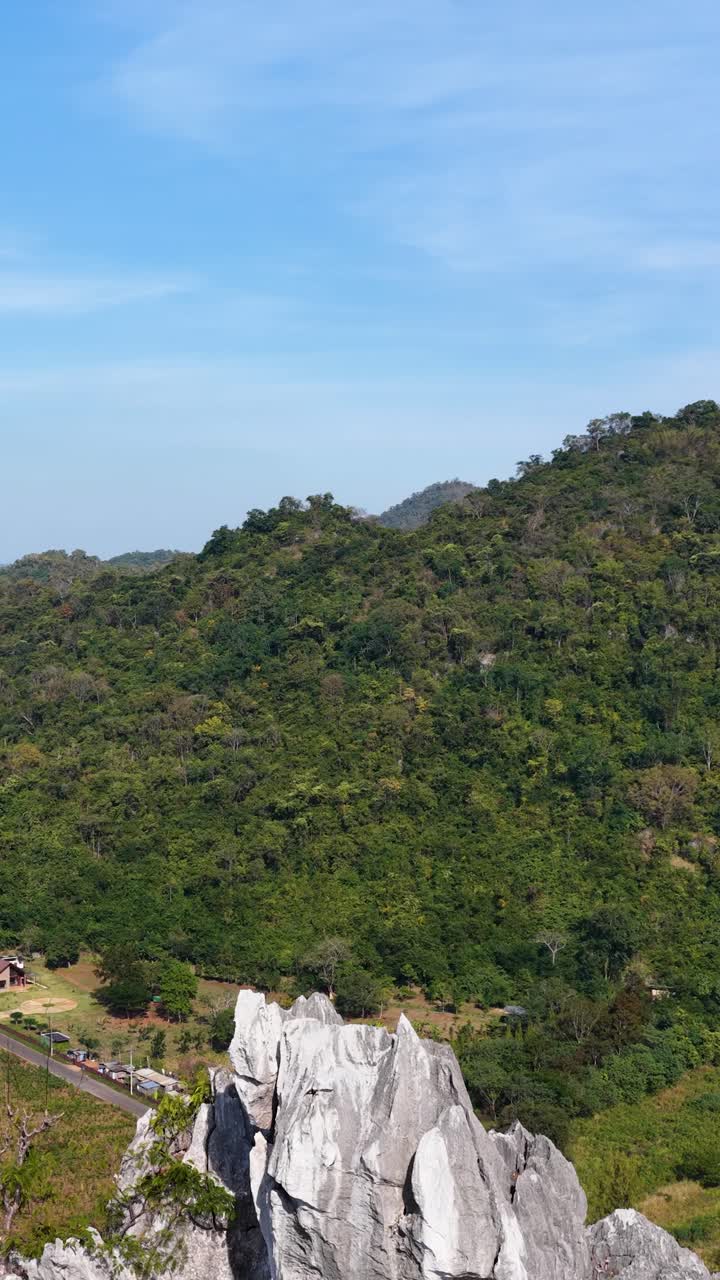 Kamera drone merekam pemandangan tebing batu kapur bergerigi yang menjulang di atas hutan hujan tropis lebat dengan langit cerah.