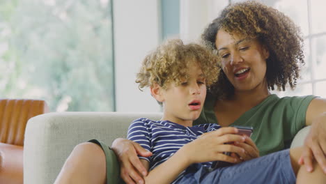 Madre-E-Hijo-Sentados-En-El-Sofá-De-Casa-Jugando-Juntos-A-Videojuegos-En-El-Teléfono-Móvil