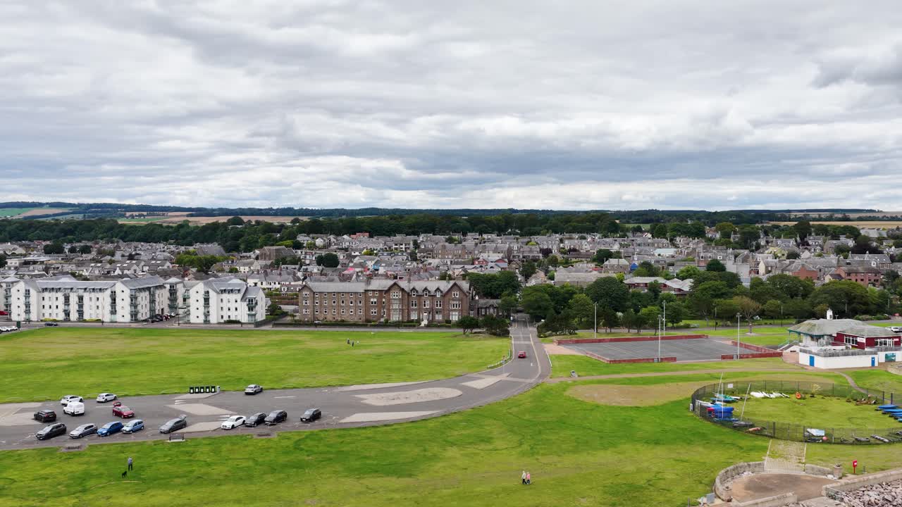 Drone ascends from beach over seawall, revealing golf course, clubhouse, and town under cloudy skies