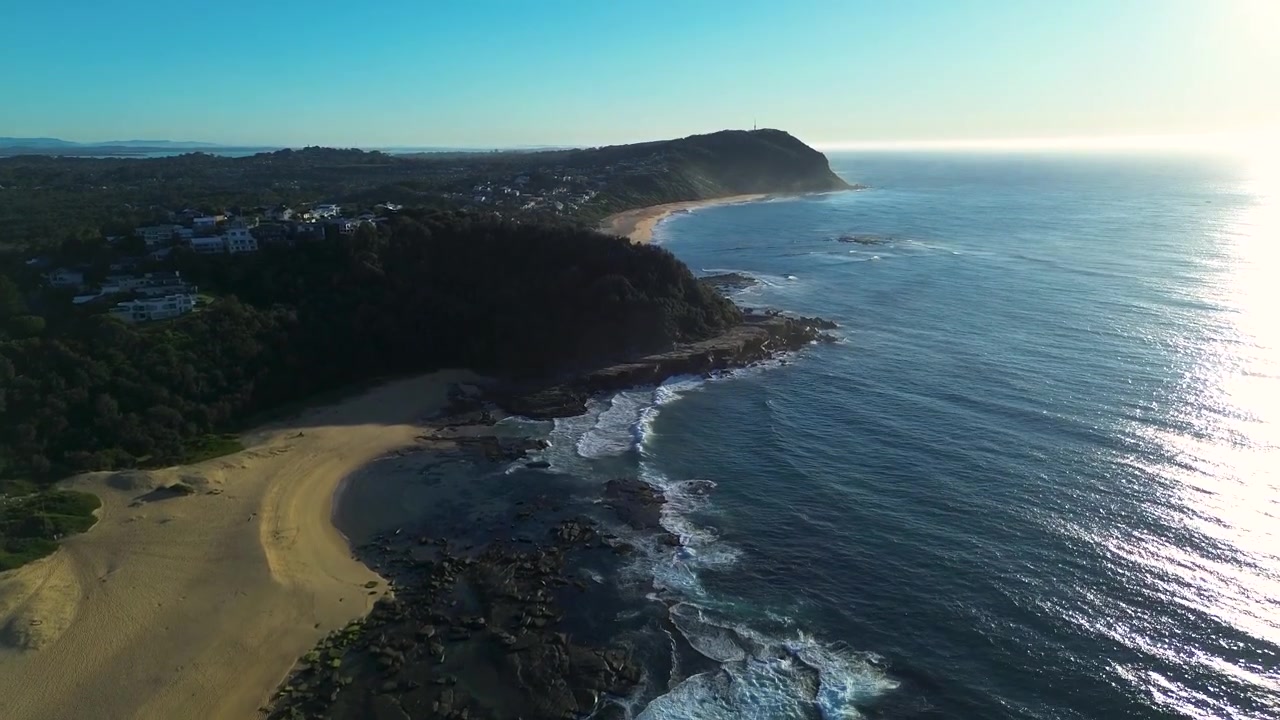 Paesaggio aereo ripreso da drone del sole scintillante che brilla sulla mareggiata dell'oceano con onde che si infrangono sulla riva di una cala sabbiosa a Spoon Bay Wamberal, con promontorio di boscaglia, Central Coast, Australia