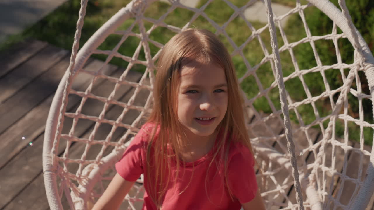 Caucasian Girl Swinging In Backyard, Red Shirt In Hanging Net Chair, Beaming With Laughter While Reaching Hands Toward Camera, Warm Sunlight On Hair, Candid Closeup Capturing Carefree Childhood Energy