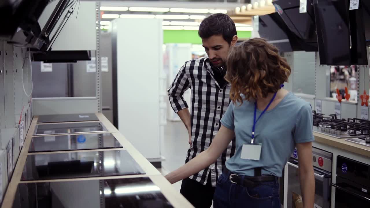 hombre de cabello oscuro, sonriente en camisa a cuadros con auriculares eligiendo una nueva cocina moderna en la tienda. él está planeando comprar muebles