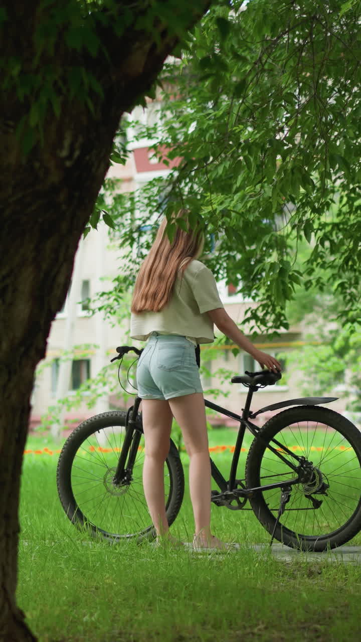 분홍색 스니커와 청바지 쇼트를 입은 젊은 여성이 나무로 둘러싸인 길에서 자전거에 접근하여 좌석에 물건을 놓고, 배경에는 울창한 초록색, 건물, 그리고 멀리 달리는 아이를 가진 사람이 있습니다.