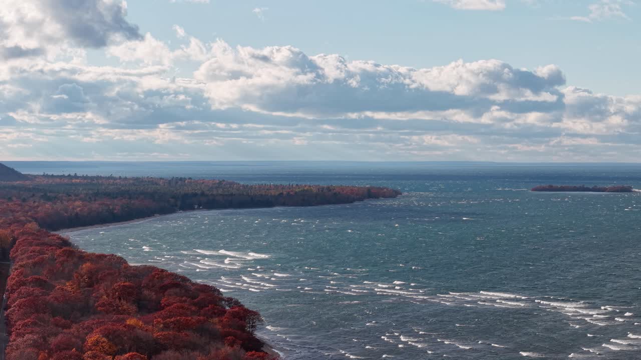 Aerial view of Lake Superior’s rugged coastline during fall, featuring vibrant red and orange forest meeting blue waters under scattered clouds