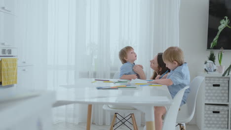 two boys brothers sit at a table in the living room and draw with pencils on paper. mom comes up and hugs the boys, smiling and stroking his head. praises and helps to make the task