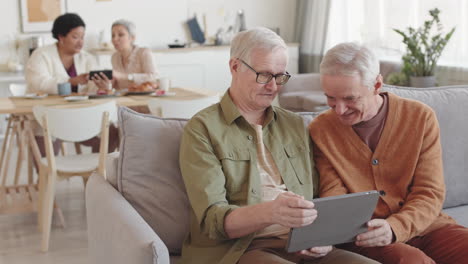 medium long of elderly caucasian male friends sitting on couch in living room, watching something on tablet computer, smiling, talking in foreground of blurred women communicating