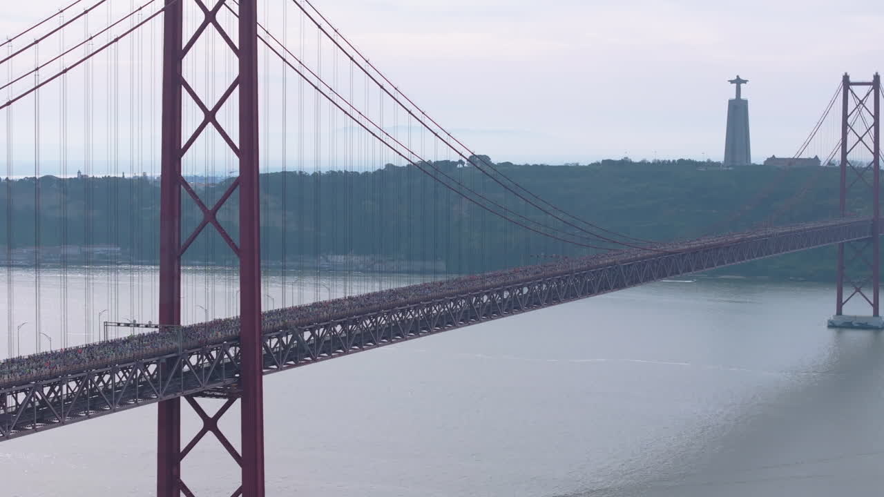 Cinematic aerial drone shot of thousands of long-distance runners crossing the iconic red 25th April suspension bridge in Lisbon, Portugal, with the Cristo Rei monument visible across the river