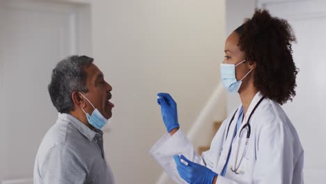 Mixed-race-female-doctor-wearing-mask-doing-swab-test-on-senior-man-at-home