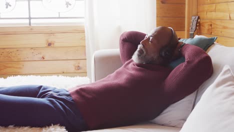 happy senior african american man in log cabin, laying on sofa and using headphones, slow motion