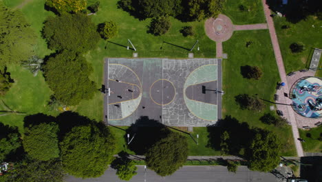 top down view of people playing basketball on concrete basketball court in outdoor park with lush green grass and trees