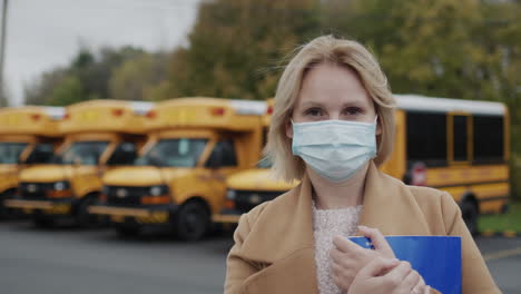 Portrait-of-a-school-teacher-in-a-protective-mask-against-the-backdrop-of-yellow-school-buses