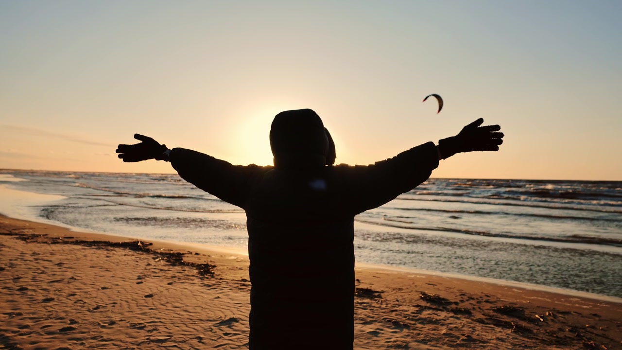 Silhouette ouvrant les bras vers un coucher de soleil doré sur la mer tandis que des kitesurfeurs s'éloignent du rivage