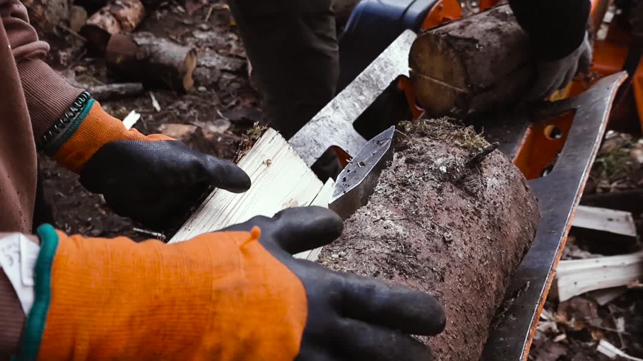Close-up of gloved hands placing logs into a wood splitter as a metal wedge splits wood into firewood pieces outdoors