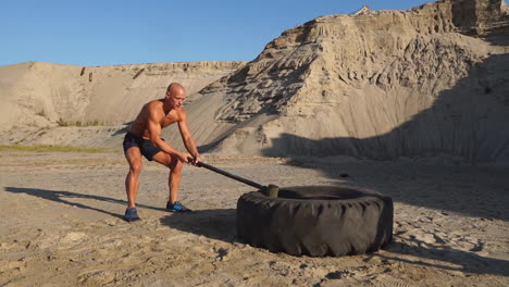 Bald-man-strongman-hits-a-hammer-on-a-huge-wheel-in-the-sandy-mountains-in-slow-motion.-strength-and-endurance-training-for-wrestlers.