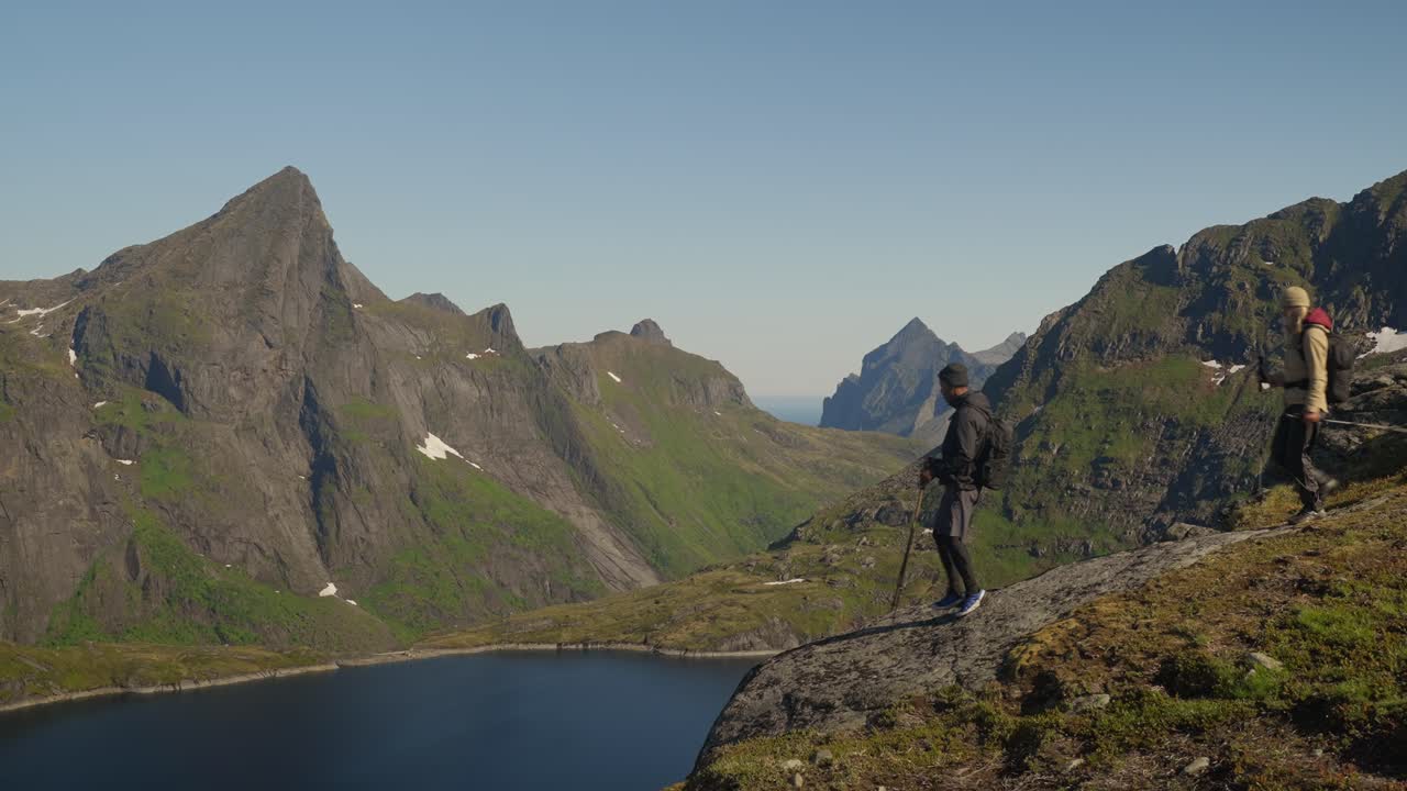 Wandelaars lopen naar een uitkijkpunt met uitzicht op een prachtig bergmeer, omringd door granieten bergtoppen, onder een helderblauwe hemel in Lofoten, Noorwegen.