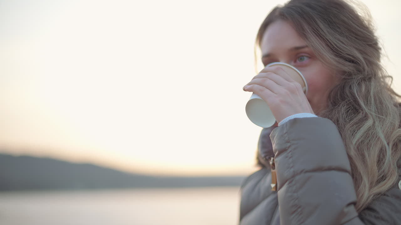 A person stands by the lake, sipping coffee from a paper cup as the sun rises. The peaceful atmosphere and gentle waves create a calm start to the day