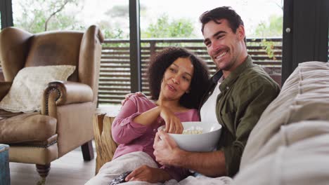 smiling mixed race couple eating popcorn while watching a movie together at vacation home