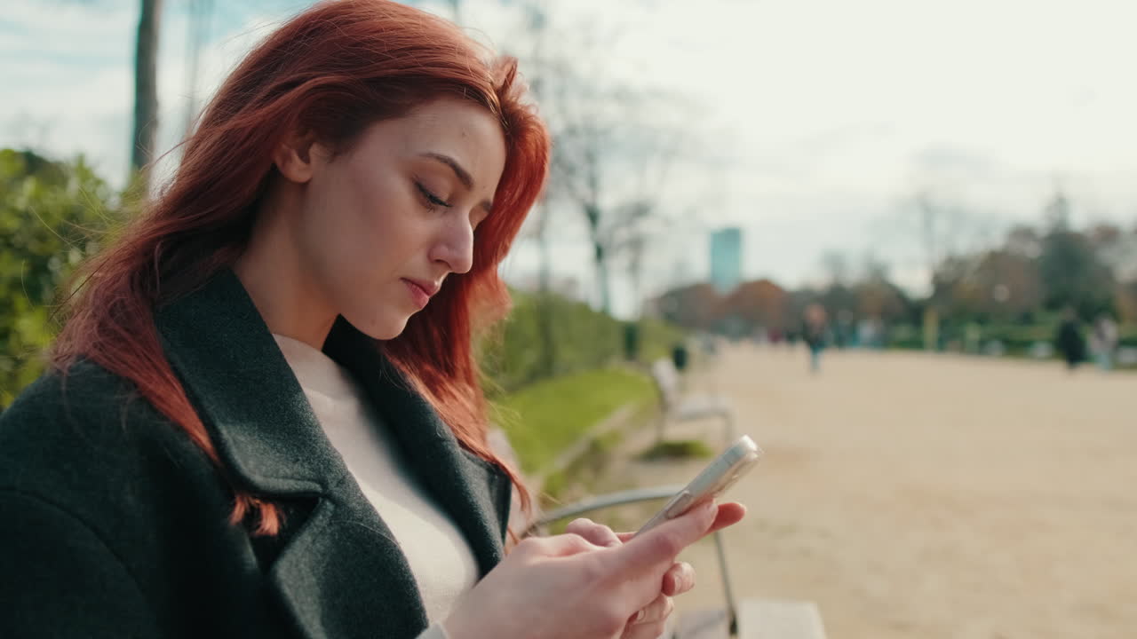 Giovane donna dai capelli rossi che usa lo smartphone in un parco