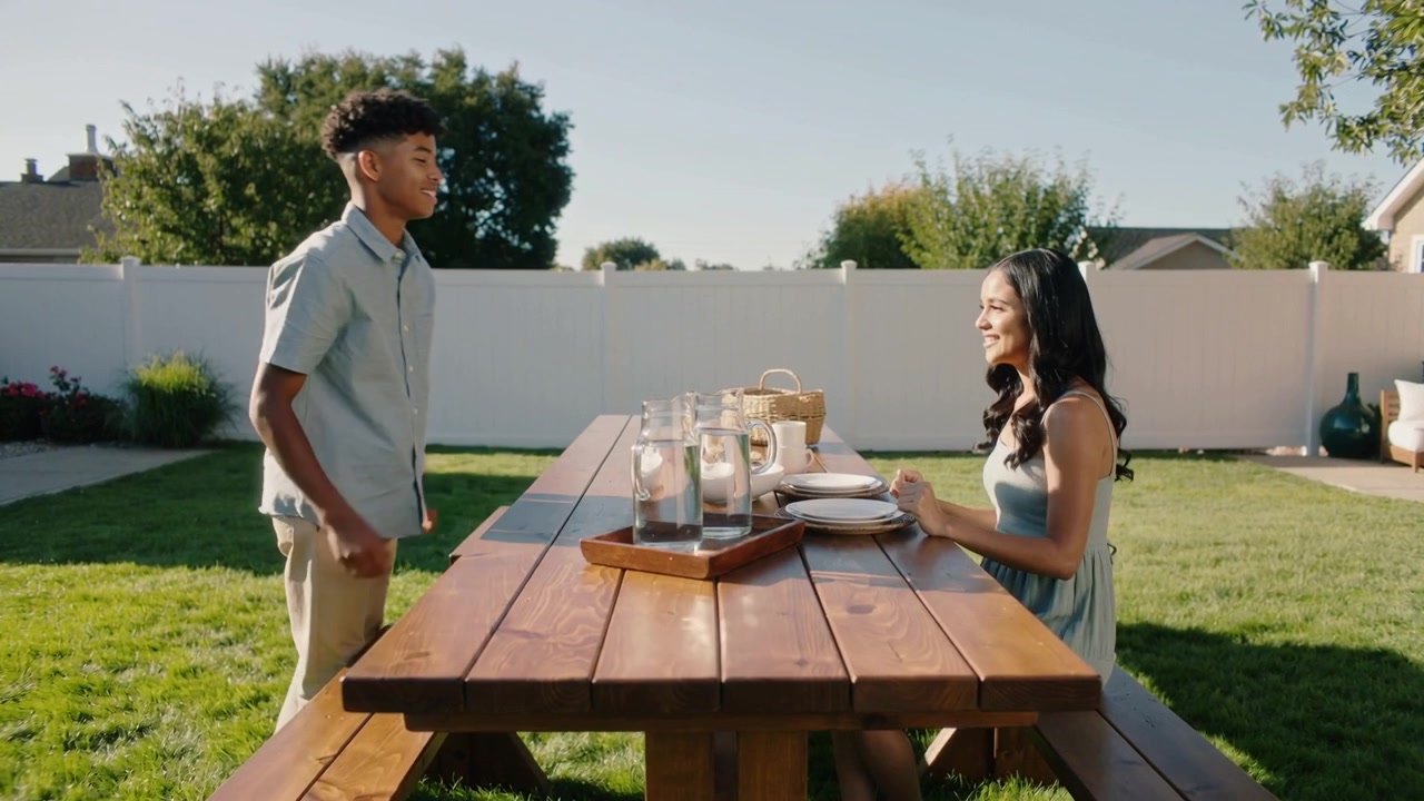 Approaching male in button-up placing three carafes on picnic table in yard to serve seated female