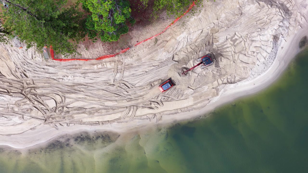 Ripresa aerea dall'alto di macchinari pesanti che spostano sabbia per la gestione costiera su una spiaggia