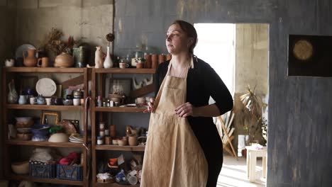 slow motion footage of young caucasian woman in casual clothes in pottery workshop, with ceramic wares and supplies on shelves and table with tools, putting on beige apron and smiling