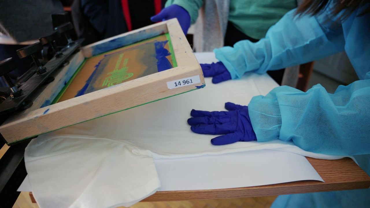 Close up footage of workers wearing protective gloves carefully positioning a white T-shirt on a screen printing press before printing a UTM logo