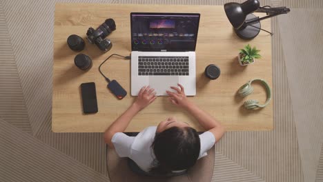 top view of a woman color grading shaking her head and having a headache while sitting in the workspace using a laptop next to the camera editing the video at home