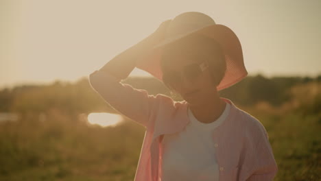 middle-aged woman wearing sunglasses and pink striped shirt over white polo, putting on sun hat during sunny day, sunlight creates warm and glowing effect, with blurred natural background