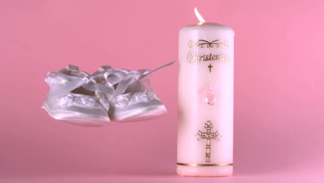 baby shoes falling beside lit baptism candle on pink background